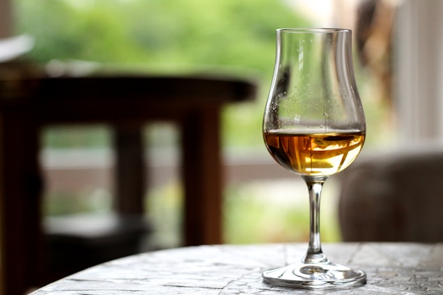 A clear glass with golden-brown liquid sits on a table with a blurred background of greenery visible through a window. The focus is on the elegant shape and reflective quality of the glass.