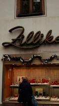 A jewelry storefront with a sign above that says 'Zillika'. The window display contains various pieces of jewelry, including necklaces and watches, arranged neatly. Above the display window, there's greenery with ornaments, suggesting a festive decoration. A person is standing in front of the window looking at the jewelry.