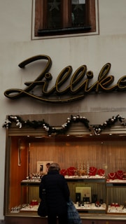 A jewelry storefront with a sign above that says 'Zillika'. The window display contains various pieces of jewelry, including necklaces and watches, arranged neatly. Above the display window, there's greenery with ornaments, suggesting a festive decoration. A person is standing in front of the window looking at the jewelry.
