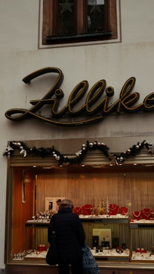 A jewelry storefront with a sign above that says 'Zillika'. The window display contains various pieces of jewelry, including necklaces and watches, arranged neatly. Above the display window, there's greenery with ornaments, suggesting a festive decoration. A person is standing in front of the window looking at the jewelry.
