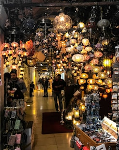 An indoor market scene with numerous colorful mosaic lamps hanging from the ceiling. The corridor is lined with various stalls displaying local handicrafts and souvenirs. Several people are walking through the market, creating a lively and bustling atmosphere. The warm glow from the lamps casts a cozy ambiance over the area.