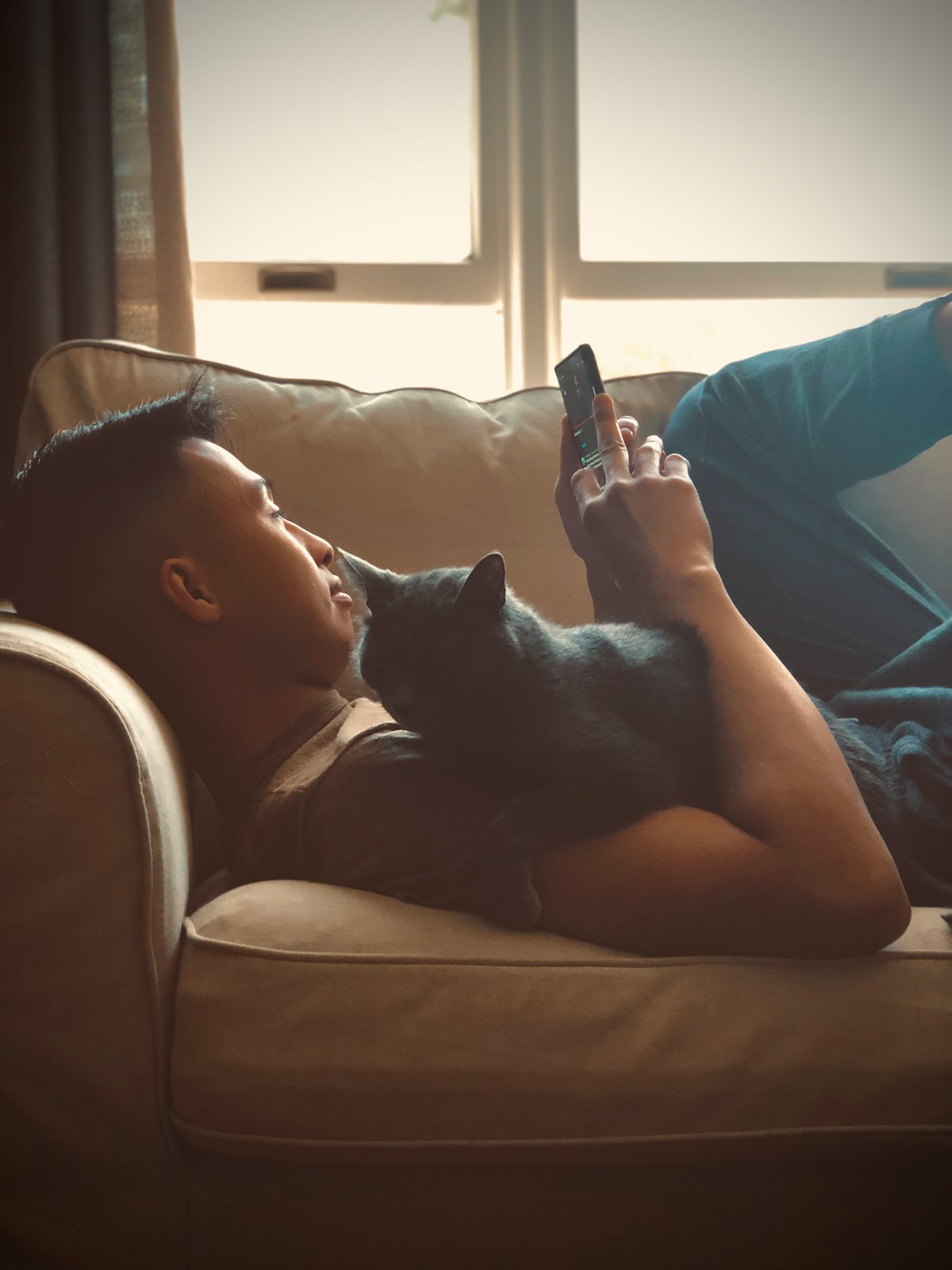 A tan man with Asian features lounging on the couch looking at his phone with a black cat resting on his chest