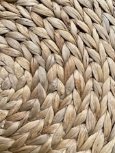 A close-up view of a tightly woven surface made of natural fibers, displaying an intricate pattern. The texture appears rough yet structured, with the fibrous strands interlaced in a repetitive and orderly manner.