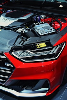 A close-up view of a car's engine bay revealing intricate components including metallic structures and cables. The car's exterior features a glossy red finish with sleek lines and a distinctive headlight design.
