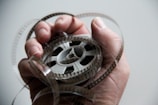 A hand is holding a roll of old-fashioned film tape with the film loosely wrapped around the fingers. The background is blurred and neutral, highlighting the focus on the film and hand.