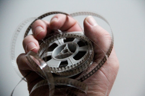 A hand is holding a roll of old-fashioned film tape with the film loosely wrapped around the fingers. The background is blurred and neutral, highlighting the focus on the film and hand.