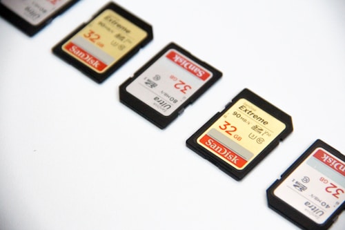 Several SD memory cards are arranged diagonally on a white surface. Each card is labeled with storage capacities and brand markings, primarily in shades of red, black, and gold.