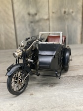 A vintage toy motorcycle with a sidecar placed on a wooden surface. The motorcycle is detailed with elements such as handlebars, a headlight, and spoked wheels. The sidecar is attached to the right side and features a metal frame and small seat, suggesting an antique design.
