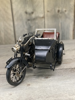 A vintage toy motorcycle with a sidecar placed on a wooden surface. The motorcycle is detailed with elements such as handlebars, a headlight, and spoked wheels. The sidecar is attached to the right side and features a metal frame and small seat, suggesting an antique design.