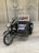 A vintage toy motorcycle with a sidecar placed on a wooden surface. The motorcycle is detailed with elements such as handlebars, a headlight, and spoked wheels. The sidecar is attached to the right side and features a metal frame and small seat, suggesting an antique design.