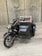 A vintage toy motorcycle with a sidecar placed on a wooden surface. The motorcycle is detailed with elements such as handlebars, a headlight, and spoked wheels. The sidecar is attached to the right side and features a metal frame and small seat, suggesting an antique design.
