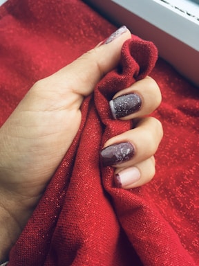 A hand with manicured nails holds a piece of red, textured fabric. The nails are painted in shades of dark purple and adorned with white decorative patterns and specks.