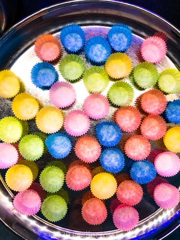 A round metal tray holds numerous colorful paper cupcake liners arranged in neat rows. The liners are vividly colored in shades of blue, pink, yellow, orange, and green, creating a bright and playful pattern against the shiny surface of the tray.