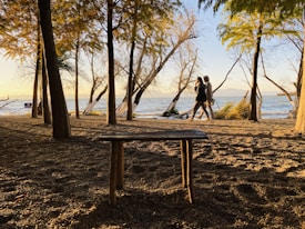 A tranquil lakeside scene with trees and a bench in the foreground. Two people are walking hand-in-hand along the shoreline in the background, with light reflecting off the water. The sun is low, casting long shadows from the trees and highlighting the pebbled ground.