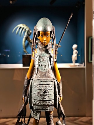 A detailed sculpture stands prominently, featuring a humanoid figure with tribal armor and elongated headgear. Intricate details in the design suggest an ancient or warrior motif. The background contains another sculptural piece with abstract, skeletal forms, creating a museum-like setting.