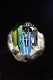 A blue and white striped jersey with the name 'Messi' and the number '10' is hanging on a line inside a room. The scene is viewed through a circular hole, giving a tunnel-like perspective. The background includes other garments and hints of patterned walls.