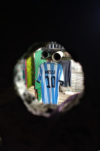 A blue and white striped jersey with the name 'Messi' and the number '10' is hanging on a line inside a room. The scene is viewed through a circular hole, giving a tunnel-like perspective. The background includes other garments and hints of patterned walls.