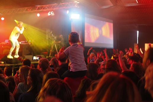 A lively concert scene with a performer on stage holding a microphone, illuminated by vibrant yellow and red stage lights. A crowd of enthusiastic attendees, including a child on someone's shoulders, watches the performance. A large screen displays colorful graphics in the background.