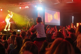A lively concert scene with a performer on stage holding a microphone, illuminated by vibrant yellow and red stage lights. A crowd of enthusiastic attendees, including a child on someone's shoulders, watches the performance. A large screen displays colorful graphics in the background.