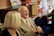 An elderly man in a suit, seated and smiling, is engaged in conversation in a cozy room filled with books and toys. A woman with blonde hair is sitting beside him, partially visible from behind. The setting appears informal and warm with shelves containing colorful toys and books in the background.