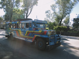 A colorful jeepney is seen driving along a road surrounded by trees. The vehicle is painted in vibrant blue, red, and yellow hues, with artistic designs covering its exterior. The street is well-lit by sunlight filtering through the trees, creating a pattern of light and shadow on the surface.