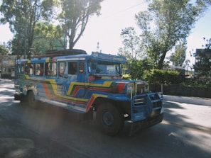 A colorful jeepney is seen driving along a road surrounded by trees. The vehicle is painted in vibrant blue, red, and yellow hues, with artistic designs covering its exterior. The street is well-lit by sunlight filtering through the trees, creating a pattern of light and shadow on the surface.
