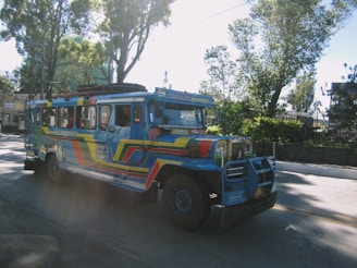 A colorful jeepney is seen driving along a road surrounded by trees. The vehicle is painted in vibrant blue, red, and yellow hues, with artistic designs covering its exterior. The street is well-lit by sunlight filtering through the trees, creating a pattern of light and shadow on the surface.