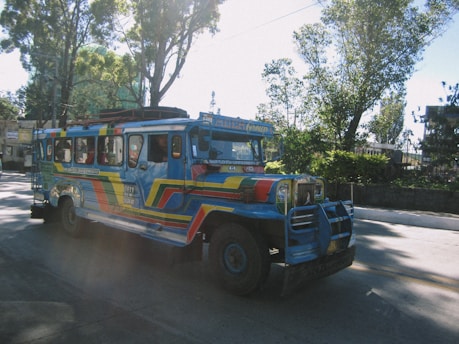 A colorful jeepney is seen driving along a road surrounded by trees. The vehicle is painted in vibrant blue, red, and yellow hues, with artistic designs covering its exterior. The street is well-lit by sunlight filtering through the trees, creating a pattern of light and shadow on the surface.