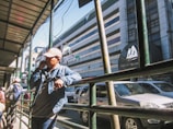 A street scene featuring a person in a denim jacket and cap talking on a cellphone while leaning on a railing. Several vehicles are parked along the roadside, and a building with the name 'Baguio Center' is visible in the background. A few pedestrians are on the sidewalk, some with backpacks.