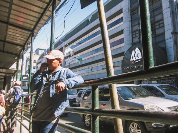 A street scene featuring a person in a denim jacket and cap talking on a cellphone while leaning on a railing. Several vehicles are parked along the roadside, and a building with the name 'Baguio Center' is visible in the background. A few pedestrians are on the sidewalk, some with backpacks.