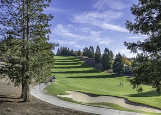 A scenic golf course featuring well-maintained, lush green fairways surrounded by tall trees under a clear blue sky. A couple of golfers are visible in the distance, near a golf cart path that curves through the landscape, and a sand trap is on the edge of the fairway.