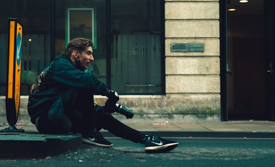 A person with medium-length hair sits on a street curb, holding a camera, and wearing dark clothing and sneakers. They appear to be engaged or focused, possibly taking a break after photography. To the left, there is a yellow traffic sign on the pavement. The background features a building with glass and stonework, partially visible interior lighting, and some windows.