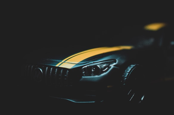 The image captures a sleek, metallic sports car with a prominent yellow racing stripe running down its dark body. The focus is on the front part of the car, highlighting its headlight and grille detail against a dimly lit background that emphasizes its contours.