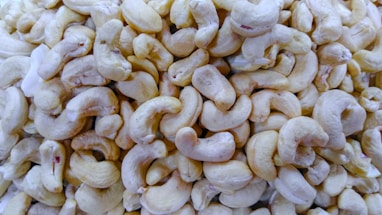 A pile of shelled cashew nuts with a creamy off-white color. The cashews are all similar in shape, with a curved, crescent-like appearance, and are densely packed together.
