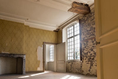A room with yellow patterned wallpaper and partially exposed plaster on the walls. A large window with multiple panes allows sunlight to stream in, illuminating the wooden floor. Part of the ceiling appears damaged or under renovation, as seen by exposed wooden beams. A decorative stone fireplace is situated against one wall without any adornments.
