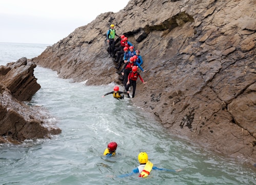 A group of people wearing helmets and protective gear are engaged in a coasteering activity along a rugged cliffside. Some participants are swimming in the water, while others are climbing or waiting on the rocky surface. The scene takes place by the sea, with overcast skies in the background.