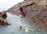 A group of people wearing helmets and protective gear are engaged in a coasteering activity along a rugged cliffside. Some participants are swimming in the water, while others are climbing or waiting on the rocky surface. The scene takes place by the sea, with overcast skies in the background.