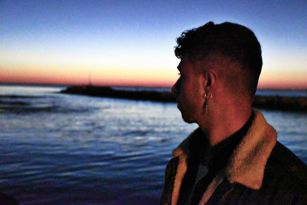 man in brown coat standing near body of water during sunset