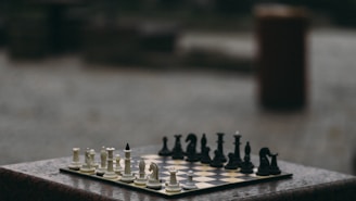 A chessboard is set up with a full array of white and black pieces arranged for a game. The setting is outdoors, possibly on a stone or marble surface, with a blurred background.