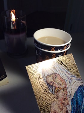 A lit candle in a glass holder casts a warm glow. Two stacked paper cups hold a light-colored liquid. A mosaic artwork with a golden background depicts a figure in a hooded robe embracing a child. The soft lighting creates a serene atmosphere.