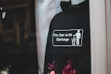 A black hoodie displayed on a mannequin, featuring a graphic with a stick figure throwing something into a trash can. The text on the hoodie reads 'Tro Dat In Da Garbage' alongside the year '2020.' There are flowers at the bottom of the image adding a pop of color.