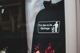 A black hoodie displayed on a mannequin, featuring a graphic with a stick figure throwing something into a trash can. The text on the hoodie reads 'Tro Dat In Da Garbage' alongside the year '2020.' There are flowers at the bottom of the image adding a pop of color.