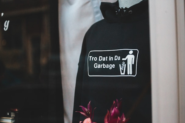 A black hoodie displayed on a mannequin, featuring a graphic with a stick figure throwing something into a trash can. The text on the hoodie reads 'Tro Dat In Da Garbage' alongside the year '2020.' There are flowers at the bottom of the image adding a pop of color.