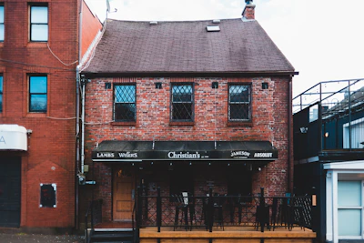 A red brick building with two stories features diamond-patterned windowpanes. The facade includes a dark awning with various brand names like Lamb's, J.P. Wiser's, and Absolut displayed. The structure has a small outdoor area with a black railing and a wooden door visible.