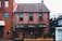 A red brick building with two stories features diamond-patterned windowpanes. The facade includes a dark awning with various brand names like Lamb's, J.P. Wiser's, and Absolut displayed. The structure has a small outdoor area with a black railing and a wooden door visible.