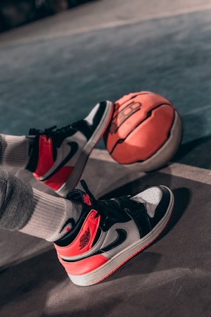A pair of stylish sneakers, featuring red, white, and black colors, is resting on a basketball. The scene is set on a sports court with a visible boundary line. The person wearing the shoes is also wearing white socks. The lighting casts a dramatic shadow on the concrete surface.