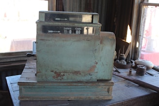 A vintage, weathered cash register sits on a wooden table in a dimly lit room. The register's surface is coated with rust and chipped paint, giving it a classic, aged look. Surrounding the register are various small objects, adding to the antique atmosphere. A window in the background allows some sunlight to filter in, highlighting the dust particles in the air.