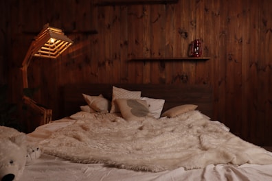 A cozy bedroom featuring a large bed with a plush, white fur blanket and several pillows arranged against a wooden headboard. There's a rustic wooden wall behind the bed with two wooden shelves, one holding a red lantern. A unique wooden floor lamp with a warm glowing light stands to the left of the bed. On the bed, there is also a stuffed polar bear toy visible.