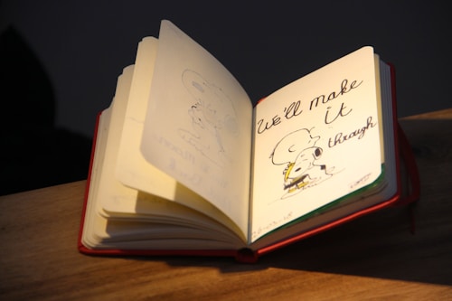 An open notebook resting on a wooden surface. The page displays an illustration of a small dog embracing a larger dog, accompanied by the handwritten text 'We'll make it through.' The surrounding environment appears dimly lit, emphasizing the page's design.