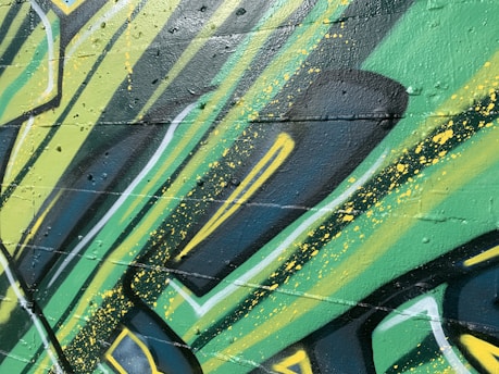 Graffiti art with bold, angular shapes and vibrant colors covers a textured wall. The design incorporates layers of green, yellow, and black with splashes of paint. The style suggests a dynamic and energetic feel, achieved through the use of sharp lines and contrasting shades.